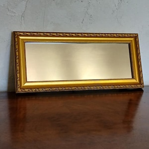 Puede incluir: Un espejo rectangular con un marco dorado y ornamentado. La superficie reflectante está dentro del marco. El espejo está sobre una superficie de madera marrón oscuro, con una pared gris claro al fondo.