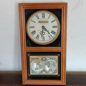 May include: A rectangular wooden framed clock with a cream-colored face and black Roman numerals. The clock face reads "Paul Masson Grande Amber Brandy". Below the clock is a framed world map.