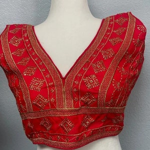 May include: A red embroidered blouse with a deep V-neckline and gold thread detailing. The embroidery features a geometric pattern.