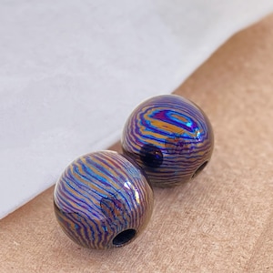 Pode incluir: Duas contas esféricas com um padrão rodopiante de azul, dourado e roxo. Cada conta tem um furo perfurado. As contas são provavelmente para fazer joias ou outros trabalhos manuais.