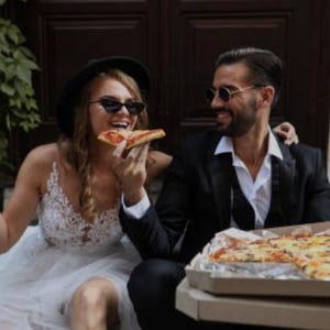 May include: A bride and groom in a relaxed setting, sharing a pizza. The bride is wearing a white dress and a black hat, while the groom is wearing a black suit and a white shirt. They are both smiling and appear to be enjoying their meal.