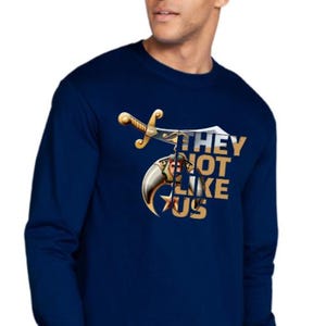 May include: Navy blue long-sleeve shirt with a graphic design featuring a sword, a crescent moon, and the text "THEY NOT LIKE US" in gold. The shirt is made of a soft material.