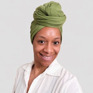 May include: A woman wearing a green head wrap. The head wrap is a solid olive green color and is wrapped in a turban style. She is wearing a white button-down shirt and smiling at the camera.
