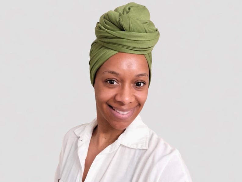 May include: A woman wearing a green head wrap. The head wrap is a solid olive green color and is wrapped in a turban style. She is wearing a white button-down shirt and smiling at the camera.