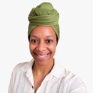May include: A woman wearing a green turban and a white button-down shirt smiles at the camera. The turban is a shade of olive green and is wrapped in a layered style. The shirt is unbuttoned at the top.