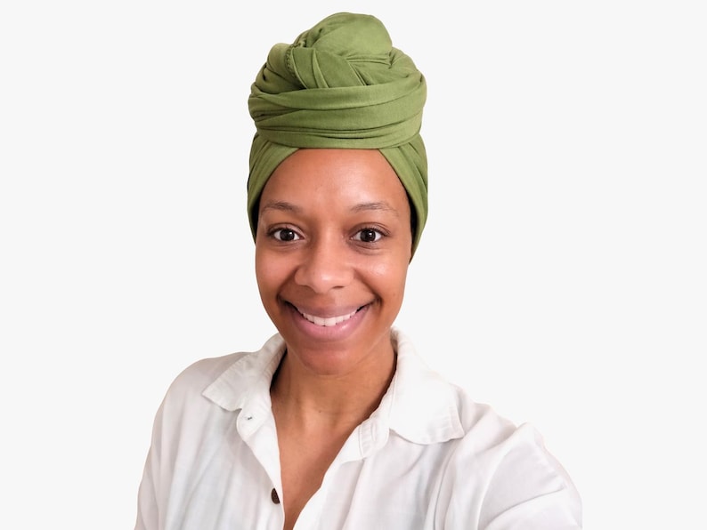 May include: A woman wearing a green turban and a white button-down shirt smiles at the camera. The turban is a shade of olive green and is wrapped in a layered style. The shirt is unbuttoned at the top.