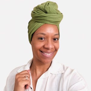 May include: A woman wearing a green turban and a white button-down shirt smiles at the camera. The turban is a solid olive green color and is wrapped in a stylish design. The woman is wearing a ring on her finger.