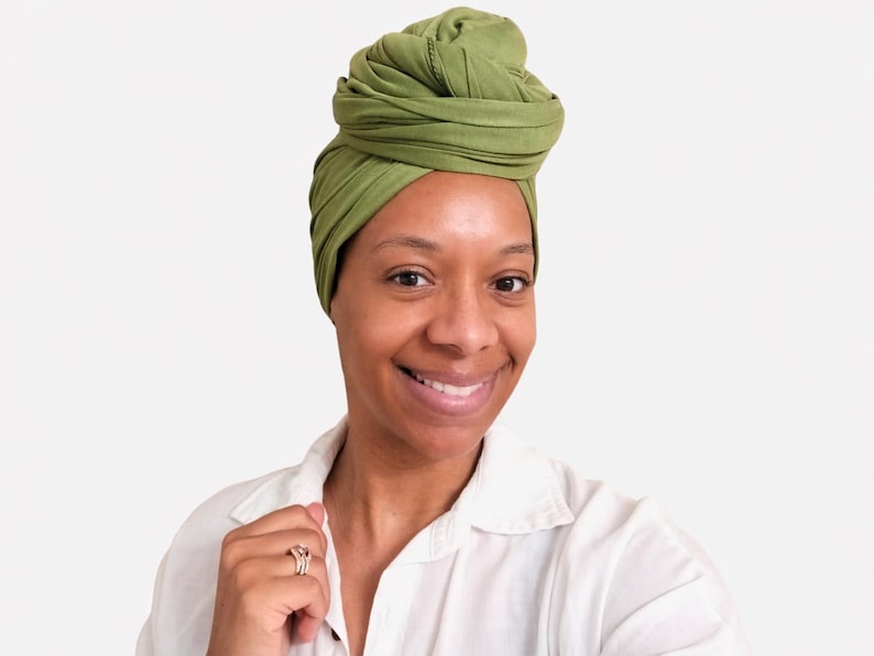 May include: A woman wearing a green turban and a white button-down shirt smiles at the camera. The turban is a solid olive green color and is wrapped in a stylish design. The woman is wearing a ring on her finger.