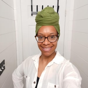 May include: A woman wearing a green head wrap and tortoise shell glasses smiles at the camera. She is wearing a white button-down shirt over a black top. The background is a white wall.