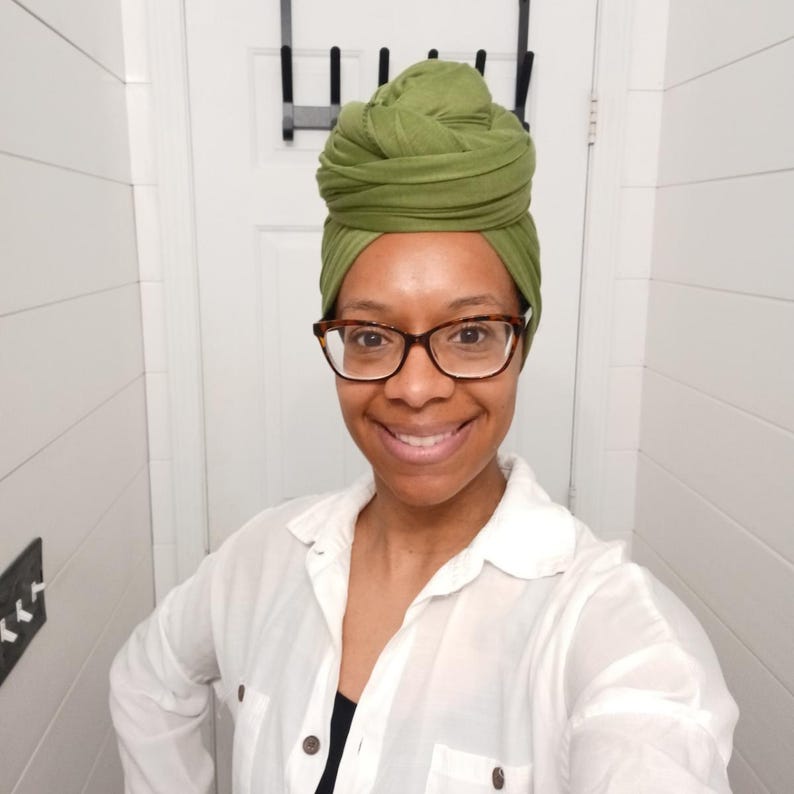 May include: A woman wearing a green head wrap and tortoise shell glasses smiles at the camera. She is wearing a white button-down shirt over a black top. The background is a white wall.