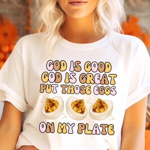 Könnte beinhalten: Weißes T-Shirt mit einem Grafikdesign, auf dem "Gott ist gut, Gott ist großartig, leg diese Eier auf meinen Teller" steht. Die Grafik ist im Retro-Stil mit braunen, rosa und gelben Buchstaben gehalten. Auf dem Teller sind drei Comic-Eier mit gelbem Dotter zu sehen.