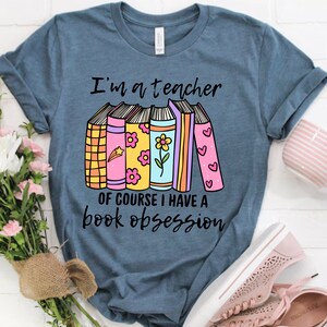 May include: A blue t-shirt with the text "I'm a teacher, of course I have a book obsession." The graphic features a stack of colorful books with floral and heart designs.