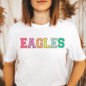 May include: White t-shirt with the word "EAGLES" in rainbow colored letters with gold glitter outlining each letter.