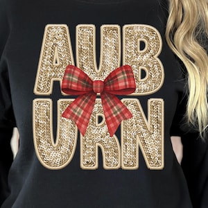 Op de afbeelding: Zwart sweatshirt met het woord "AUBURN" in gouden pailletten. Een rood-groene geruite strik is gecentreerd over de letters. De sweatshirt heeft een ronde hals.