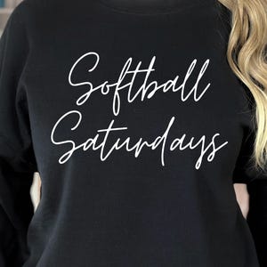 May include: Black sweatshirt with the white cursive text "Softball Saturdays". The sweatshirt is a crew neck style, and the text is centered on the front. The image shows a close-up of the upper body.