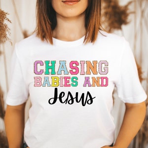 Może przedstawiać: Biała koszulka z napisem "CHASING BABIES AND Jesus" w kolorowych literach. Tekst jest w zabawnym, swobodnym kroju pisma.