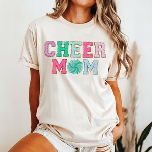 Könnte beinhalten: Ein beiges T-Shirt mit dem Text "CHEER MOM" in bunten Glitzerbuchstaben und einem blauen und grünen Pompon-Motiv.