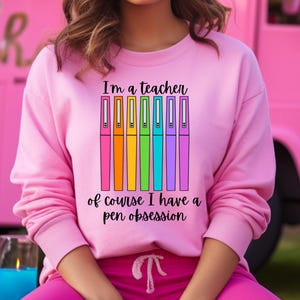 May include: Pink sweatshirt with the text "I'm a teacher of course I have a pen obsession" and a rainbow of pens.