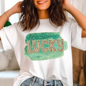 Könnte beinhalten: Weißes T-Shirt mit einem grünen und goldenen Glitzerdesign, auf dem "LUCKY" steht.