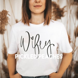 May include: White t-shirt with the text "Wife" in a cursive font with a small heart above the "i". Below the text is the phrase "PICKLED PEACHES".