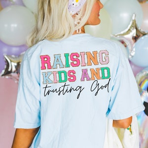 May include: Light blue t-shirt with the text "Raising Kids and Trusting God" in colorful letters with gold glitter accents.