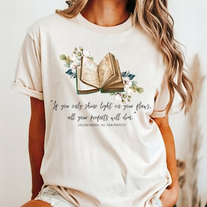 May include: A beige t-shirt with a floral design and an open book. The text on the shirt reads "If you only shine light on your flaws, all your perfects will dim." -Colleen Hoover, "All Your Perfects"