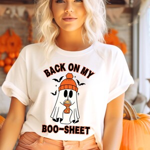 May include: White t-shirt with a Halloween-themed graphic. The design features a ghost wearing an orange beanie and holding a drink, with the text "BACK ON MY BOO-SHEET" in orange and black. Bats are also included in the design.