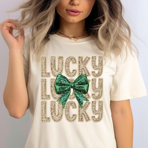 Könnte beinhalten: Ein weißes T-Shirt mit dem Wort "LUCKY" dreimal in goldenen Glitzerbuchstaben wiederholt. Eine grüne Glitzer Schleife befindet sich mittig zwischen den Wörtern.