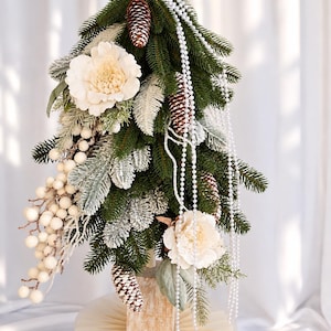 May include: A tabletop Christmas tree featuring green pine branches, frosted foliage, brown pinecones, and large cream-colored flowers. The tree is decorated with long strands of white faux pearls and clusters of small white berries, set on a wooden base.