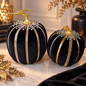 May include: Two black velvet pumpkins featuring gold stems and vertical decorative strips. The pumpkins are adorned with rows of clear, reflective rhinestones and metallic gold accents that extend from the top stems down the sides.
