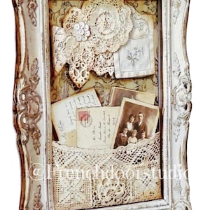 May include: Ornate, white framed shadow box with vintage lace, old photographs, and handwritten letters. The frame has a distressed finish, and the interior is lined with patterned paper. A crocheted pocket holds some of the ephemera.