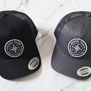 May include: Two snapback trucker hats, one black and one charcoal grey, featuring mesh back panels and a circular patch on the front. The patch displays a compass rose graphic with the text Riverside Traders Canadian Wilderness Goods.
