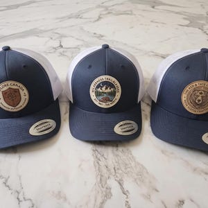 May include: Three navy blue and white snapback caps featuring circular leather patches on the front. The left patch shows a shield and compass, the centre patch depicts a mountain landscape with a beaver, and the right patch displays a bear head.