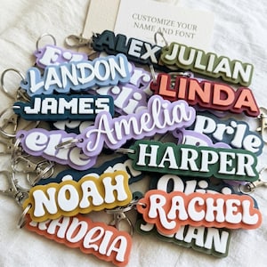 Peut inclure: Une collection de porte-clés personnalisés affichant divers prénoms comme Alex, Julian, Landon, Linda, James, Amelia, Harper, Noah et Rachel dans des polices et couleurs variées, chacun muni d'un mousqueton et d'un anneau en métal argenté.