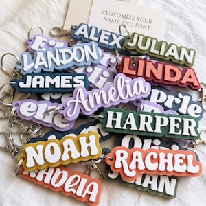 May include: A collection of personalised keychains featuring various names like Alex, Julian, Landon, Linda, James, Amelia, Harper, Noah, and Rachel. Each keychain has a layered design with white raised lettering on colourful, shaped plastic bases.