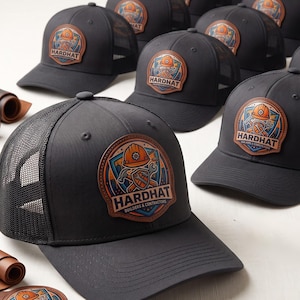 May include: A collection of black trucker hats featuring a circular leather patch on the front. The patch displays an orange hard hat, crossed tools, and the text HARDHAT BUILDERS & CONTRACTORS in a shield design with blue and orange accents.