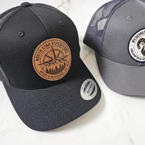 May include: A black mesh-back snapback cap with a tan leather patch featuring a compass and mountain graphic labelled North Star Expeditions, alongside a grey mesh-back cap with a circular patch depicting a ram and Wilderness Trailblazers text.