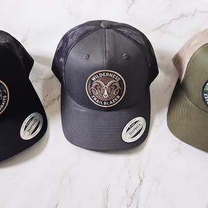 May include: Three trucker caps with mesh backs and snapback fastenings. The black cap features a mountain patch with the text Adventure Awaits. The charcoal cap has a ram patch with Wilderness Trailblazer text. The olive cap shows an eagle patch with Explore Further text.