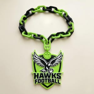 May include: A necklace featuring a chain of alternating black and lime green plastic links. A large shield-shaped pendant hangs from the chain, displaying a black and white eagle graphic, a star icon, and the text HAWKS FOOTBALL in black letters.