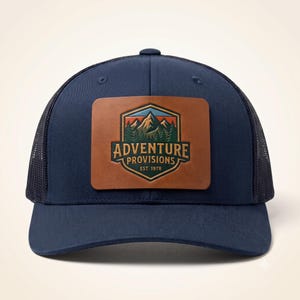 Peut inclure: Une casquette trucker bleu marine avec un écusson rectangulaire en cuir marron sur le devant. L'écusson présente un motif de montagne et de forêt avec le texte ADVENTURE PROVISIONS EST. 1978 dans un emblème en forme de bouclier aux tons orange et bleu.