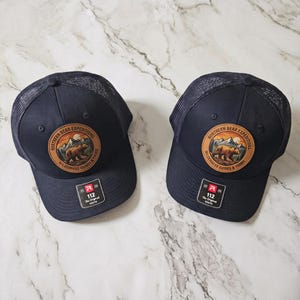 May include: Two navy blue trucker caps with mesh backs and adjustable snapback closures. Each cap features a circular leather patch with a graphic of a bear in a mountain landscape and the text Northern Bear Expeditions Wilderness Guides & Tours.