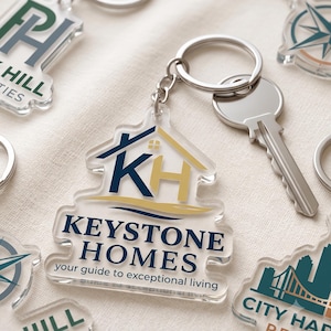 Peut inclure: Un porte-clés en acrylique transparent en forme de maison, arborant un logo bleu foncé et doré avec les lettres KH et le texte KEYSTONE HOMES your guide to exceptional living. Un anneau en métal argenté est fixé avec une clé argentée.