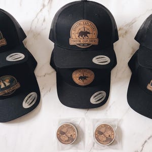 May include: Five black snapback caps featuring circular leather-look patches with Coastal Explorers text and nature-themed graphics, including a sailing boat, a bear, and an orca. Two small, round, matching patches are packaged in clear plastic bags below.