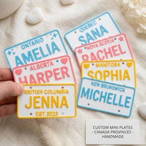 May include: A collection of rectangular white mini license plates featuring various Canadian provinces and names. The plates include Ontario with Amelia, Quebec with Sana, Nova Scotia with Rachel, Alberta with Harper, Manitoba with Sophia, New Brunswick with Michelle, and British Columbia with Jenna. Each plate has coloured borders and text.