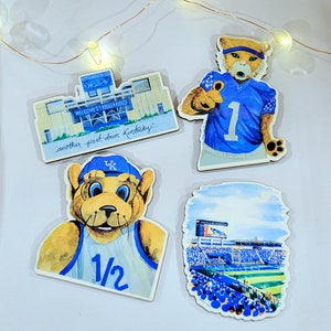 May include: Four wooden magnets featuring a blue and white mascot with the text "1/2" and "UK", a blue and white mascot with the text "1", a stadium with the text "Welcome to Kroger Field" and "another first down Kentucky", and a stadium with a crowd of people.