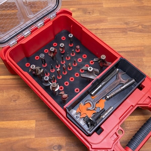 May include: A red plastic tool storage case with a clear lid containing a black foam insert with multiple circular cutouts holding various metal router bits, along with a separate black tray containing a metal wrench and additional tools.