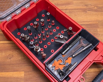 May include: A red plastic tool storage case with a clear lid containing a black foam insert with multiple circular cutouts holding various metal router bits, along with a separate black tray containing a metal wrench and additional tools.