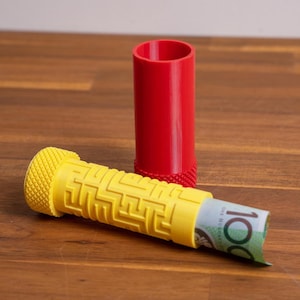 May include: A cylindrical red container with a textured base and a yellow cylindrical puzzle case featuring a maze pattern on the exterior. The yellow case is partially open, revealing a rolled-up one hundred dollar bill tucked inside the hollow centre.