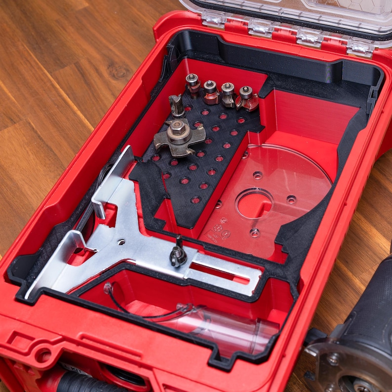 May include: A red plastic storage case with a clear lid containing a black foam insert. The insert holds a metal router guide, a clear circular base plate, and several small metal router bits arranged in a grid pattern within the case.