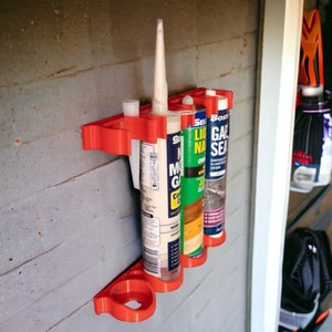 Puede incluir: Un organizador de pared de plástico naranja brillante que sostiene tres tubos verticales de adhesivo de construcción y sellador. El soporte cuenta con recortes circulares para la base de cada tubo y un marco superior para mantenerlos rectos.